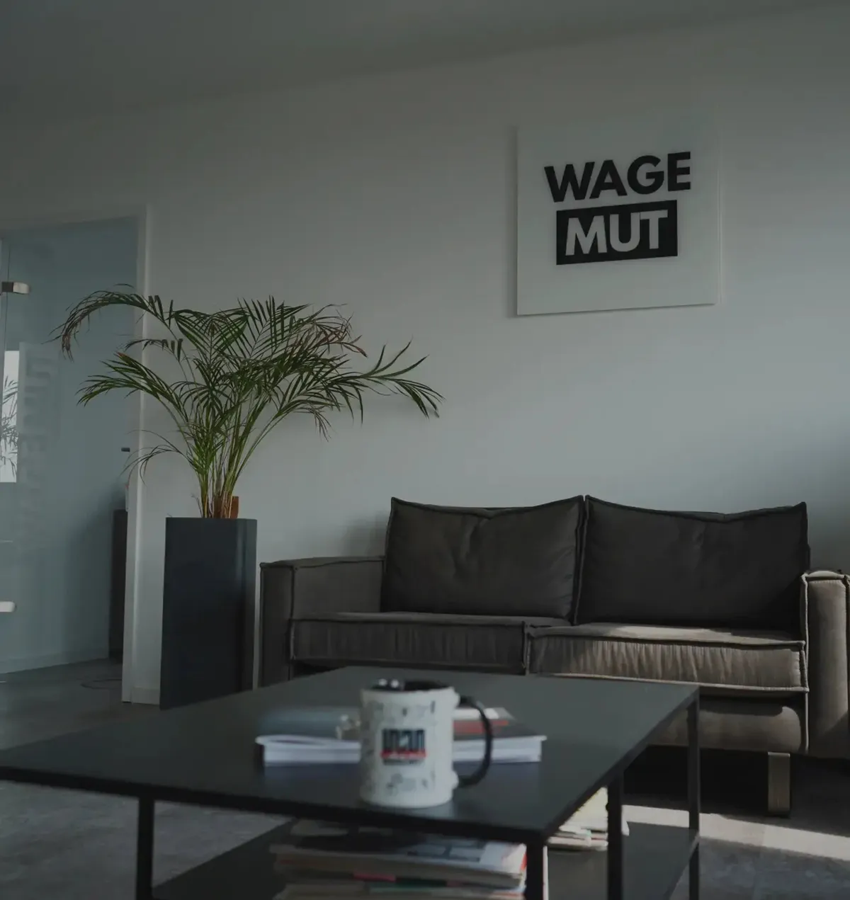 Lounge at the WAGEMUT studio in Düsseldorf: dark sofa, coffee table with a mug, a plant and the WAGEMUT lettering on the wall