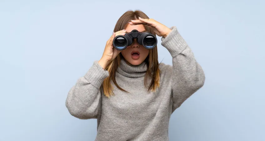 Frau im grauen Rollkragenpullover schaut überrascht durch ein Fernglas – selektive Wahrnehmung im Marketing